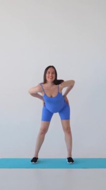 Beyaz arka planda fitness eğitimi veren, kalçalarını esneten orta yaşlı, pozitif beyaz bir kadının portresi. Dikey video.