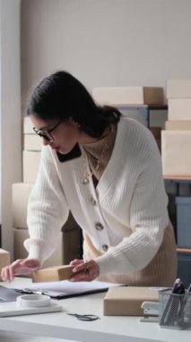 Diz üstü bilgisayarla çalışan ve depo ofisinde telefonla müşteriyle konuşan neşeli, genç, beyaz bir iş kadını. Dikey video.