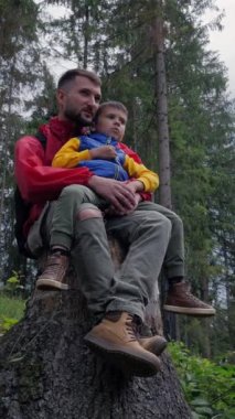 Okul çocuğu ve babası tatil sırasında yemyeşil bir ormanda dinlenerek birlikte sakin bir anın tadını çıkarıyorlar. Doğa gezisinin kıymetini bilirken aile bağı kurmak. Dikey video.