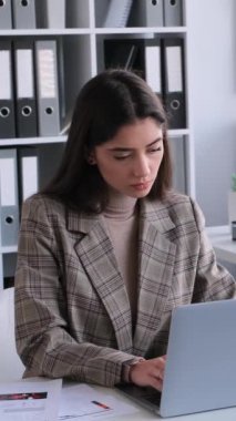 Ofiste dizüstü bilgisayarla çalışan ve iş arkadaşıyla sohbet eden neşeli, beyaz, genç bir iş kadını. Strateji analizi. Dikey video.