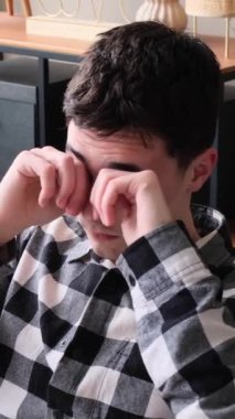 Hüzünlü ve sağlıksız genç beyaz bir adam gözlerini ovuşturuyor ve evde esniyor. Aşırı gerilim, yorgunluk ve stres kavramı. Dikey video.