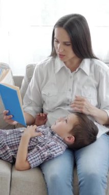 Neşeli orta yaşlı beyaz anne ve oğlu boş zamanlarını evde geçirip kanepede dinlenirken kitap okuyorlar. Dikey video.