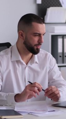 Konsantre Kafkasyalı orta yaşlı işadamı çevrimiçi video görüşmeleri ve bürodaki belgelerle çalışıyor. İş iletişimi kavramı. Dikey video.
