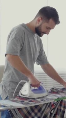 Orta yaşlı, beyaz bir adam evde kıyafetlerini ütülüyor. Günlük ev işleri ve ev işleri konsepti. Dikey video.