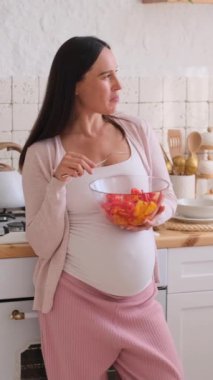 Neşeli orta yaşlı, beyaz hamile kadın mutfakta sağlıklı yiyecekler yiyor. Hamilelik ve taze yemek kavramı. Dikey video.