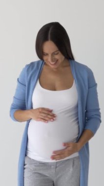 Mutlu hamile kadın beyaz bir arka planda duruyor, karnına özenle dokunuyor, sevinç, beklenti ve bebek için aşkı ifade ediyor. Dikey video.