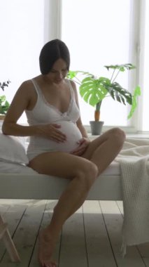 Dost canlısı hamile bir kadın yatak odasında karnını ovalıyor, doğum için sevgi ve beklenti gösteriyor. Sağlık, bağlılık ve duygusal bağ. Dikey video.