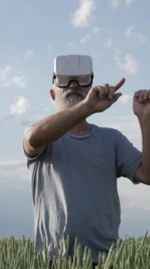 Yaşlı bir adam tarlada duruyor, VR kulaklığı kullanıyor. Doğadaki modern teknoloji, açık hava ortamındaki gelecekteki dijital deneyimler. Dikey video.