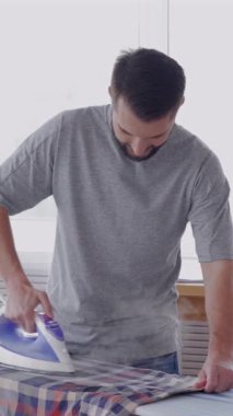 Keyifli Kafkasyalı orta yaşlı bir adam evde gömlek ütülüyor. Her gün sabah rutini ve temizlik konsepti. Dikey video.