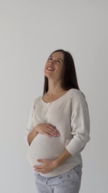 Beyaz arka planda karnını okşayan gülümseyen hamile bir kadın. Sevgi, şefkat, neşeli bir beklenti ve doğum bekliyor. Dikey video.