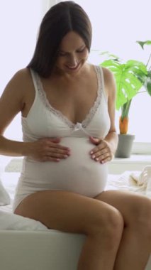 Mutlu hamile bir kadın evde yatağında oturuyor, elleriyle karnına dokunuyor, mutluluğu ve bebek beklentisini ifade ediyor. Gelecekteki ebeveynlik ve duygusal bağ. Dikey video.