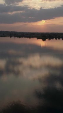 Ormanla çevrili bir gölün ya da gölün hava görüntüsü, günbatımının canlı renkleriyle akşam gökyüzünü doğal bir vahşi yaşam ortamında resmediyor. Dikey video.