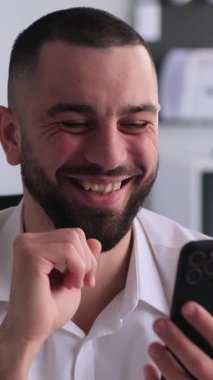 Arkadaş canlısı, beyaz, orta yaşlı bir adam ofiste çalışırken online eğlence için akıllı telefon kullanıyor. Sanal alemde boş zaman kavramı. Dikey video.