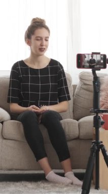 Mutlu kadın çevrimiçi sosyal medya izleyicileri için video kaydediyor, takipçilerle konuşurken evdeki bir kanepede rahatça oturuyor. Dikey video.