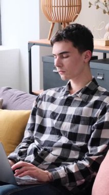 Düşünceli Kafkasyalı genç adam dizüstü bilgisayar kullanıyor, internette çalışıyor ya da internette geziniyor, evdeki oturma odasındaki kanepede oturuyor. Dikey video.