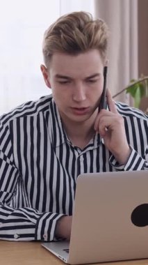 İş adamı laptopta çalışırken aynı zamanda modern ofis ortamında telefonla konuşuyor, iş ve iş iletişimi arasında birden fazla iş yapıyor. Dikey video.