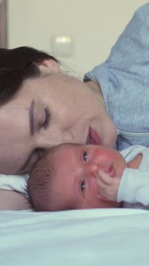 Şefkatli anne yeni doğmuş bebeği kucaklar ve öper, evde oturma odasında yatakta dinlenirken. Annelik ve ebeveynlik kavramı. Dikey video.