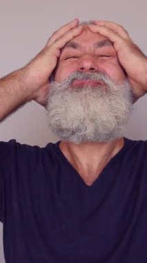 Beyaz arka planda dikilerek baş ağrısından muzdarip Kafkasyalı kıdemli bir adam. Yorgunluk, hayal kırıklığı ve keder konsepti. Dikey video.