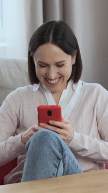 Neşeli bir kadın internette geziniyor ve evde çevrimiçi boş vakitlerin tadını çıkarıyor. Telefonunu eğlence ve iletişim için kullanıyor ve dinleniyor. Dikey video.