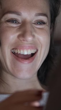 Akşamları internet gezintisi, iletişim ve dinlenmenin tadını çıkarırken akıllı telefonu evde eğlenmek için kullanan heyecanlı bir kadın. Dikey video.