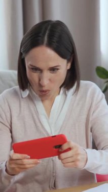 Neşeli bir kadın telefonda video oyunları oynuyor, online yarışmalara katılıyor ya da evde boş vakitlerin tadını çıkarıyor. Oyuna dalmış, eğleniyor ve dijital eğlenceye dalmış. Dikey video.