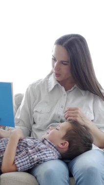Neşeli küçük çocuk ve beyaz orta yaşlı anne birlikte kitap okuyor boş zamanlarını evde kanepede dinlenerek geçiriyorlar. Dikey video.