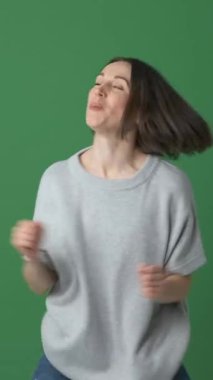 Yeşil arka planda dans ve müzikten hoşlanan aktif bir kadın, ritmik olarak hareket ederken zevk, heyecan ve enerji hissediyor, eğlence ve hareketliliği kucaklıyor. Dikey video.