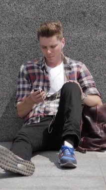 Öğrenci dışarıda dinleniyor, internetten mesajlaşıyor ve internet eğlencesi için telefon kullanıyor. Boş zamanın tadını çıkarmak, teknolojiyle rahatlamayı birleştirmek. Dikey video.