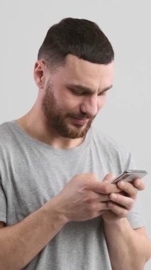 Olumlu, akıllı telefonuyla internet keyfi yapan beyaz arka planda mesajlaşırken gülen bir adam. İletişim ve eğlence rahat bir anda yakalanır. Dikey video.