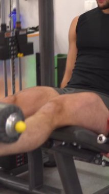 Spor antrenmanı ve spor salonunda spor yaparken bir makinede bacak uzatma egzersizi yapan kararlı bir sporcu. Güç ve odaklanma. Dikey video.