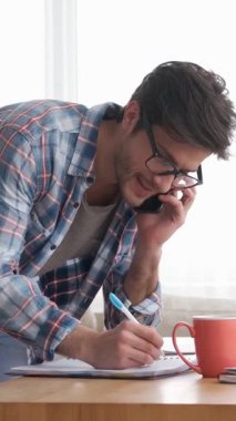 Odaklanmış uzaktan çalışan evden çalışırken iş görüşmesi için cep telefonu kullanır. Bağlantılı ve konuşkan kalıyor. Dikey video.