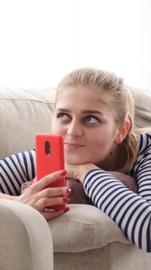 Dost canlısı bir kadın evde cep telefonuyla rahatlıyor, internet ve internet iletişiminin tadını çıkarıyor. Dijital eğlencenin rahatlığı. Dikey video.