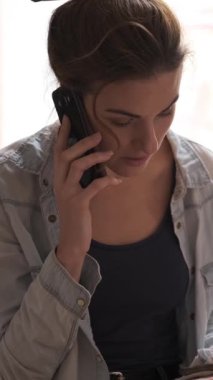 Olumlu kadın, bir sanat atölyesinde resim yaparken telefonla konuşuyor, yaratıcılıkla iletişimi birleştiriyor. Dikey video.