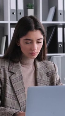 Düşünceli, genç, beyaz kadın, dizüstü bilgisayarla çalışıyor ve ofiste profesyonel muhasebe işleriyle uğraşıyor. Dikey video.