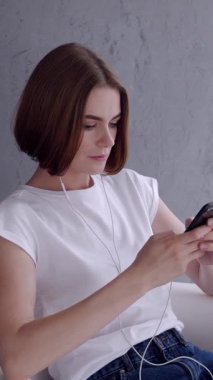 Rahat kadın, gri arka planda cep telefonu kullanarak internette boş zaman geçiriyor. İletişim, rahatlama ve dijital bağlantı. Dikey video.