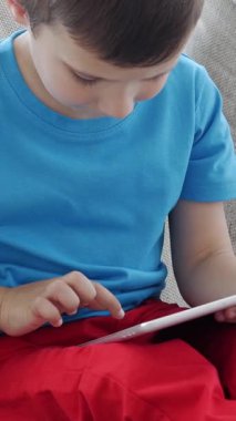 Tablet PC kullanan neşeli küçük beyaz çocuk boş zamanlarını evde internette geçiriyor. Çevrimiçi eğitim ya da eğlence anlayışı. Dikey video.