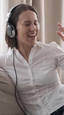 Mutlu kadın evdeki kanepede oturmuş, müzik dinlerken eşlik ediyor. Eğlencenin ve ferahlığın tadını çıkarıyor, neşeli ve canlandırıcı atmosferi tamamen kucaklıyor. Dikey video.