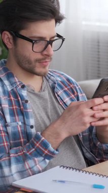 Evde çalışan düşünceli bir çalışan, online sohbet için telefon kullanıyor. İşletme ve teknolojiye odaklanmış, görevleri uzaktan hallederken bağlı kalıyor. Dikey video.