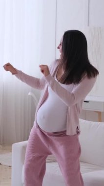 Heyecanlı ve aktif Kafkasyalı orta yaşlı hamile kadın müzik eşliğinde dans ediyor ve evde eğleniyor. Dikey video.