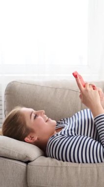 Evdeki kanepede dinlenirken akıllı telefon kullanan neşeli kadın mesajları. Dinlenme zamanlarında internette sohbet ve internet eğlencesi. Dikey video.
