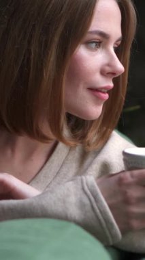 Huzurlu kadın evdeki kanepede dinlenip düşünürken çay içiyor. Zevk, rahatlık ve dinlenme anları. Dikey video.