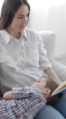 Kafkasyalı orta yaşlı anne ve küçük okul çocuğu birlikte kitap okuyup, ailecek evde vakit geçiriyorlar. Dikey video.