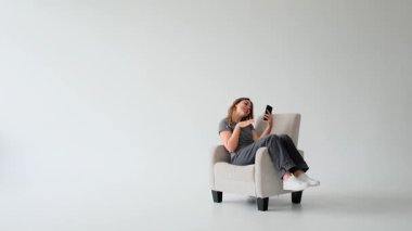 Heyecanlı Kafkasyalı genç bir kadın telefonuyla görüntülü konuşma yapıyor, beyaz arka planda koltukta dinleniyor. Sanal sohbet ve iletişim.