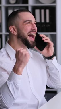 Modern ofiste çalışırken iş görüşmeleri sırasında başarıyı kutlayan arkadaş canlısı, orta yaşlı beyaz bir adam. Dikey video.