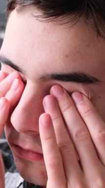 Evde oturma odasındaki kanepede gözlerini ovuşturan sinirli beyaz bir gencin yakın portresi. Yorgunluk ve aşırı yükleme kavramı. Dikey video.