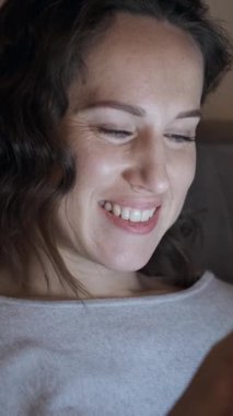 Gülümseyen bir kadın akşamları evde telefonuyla eğleniyor, internette geziniyor ve uzun bir günün ardından dinleniyor. Dikey video.