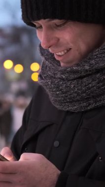 Neşeli adam dışarıda dolaşıyor, cep telefonuyla sosyal medyada mesajlaşıyor. Şehir ortamında boş zaman ve dijital eğlence. Dikey video.