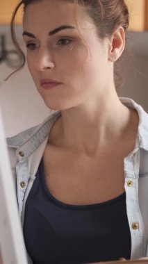 Stüdyoda resim yapmak için boya fırçası, palet ve yağ boyası kullanan, sehpadaki tuvale odaklanmış bir kadın. Dikey video.