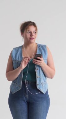 Şık ve neşeli bir kadın dans ediyor ve elinde telefon, beyaz bir arka planda şarkı söylüyor, eğlencenin tadını çıkarıyor. Dikey video.