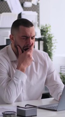 Uykulu, beyaz, orta yaşlı bir çalışan ofiste çalışırken esniyor ve esniyor. Aşırı çalışma ve hayal kırıklığı kavramı. Dikey video.